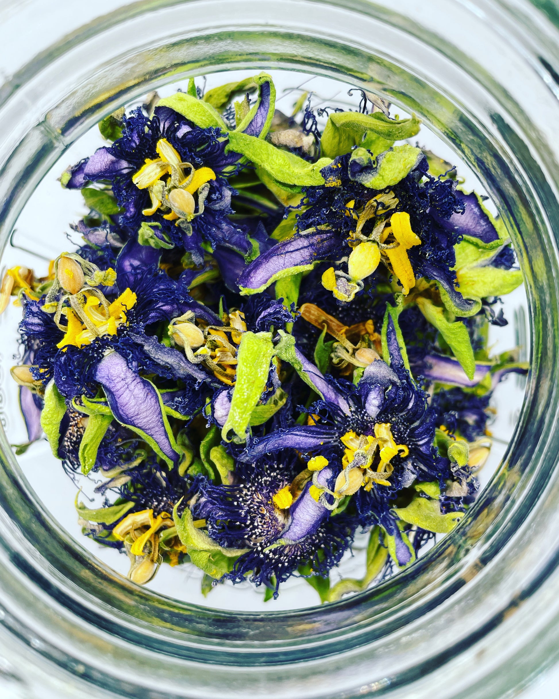 Dried passionflower blossoms (Passiflora incarnata) with purple petals, green sepals, and yellow stamens stored in a clear glass jar.